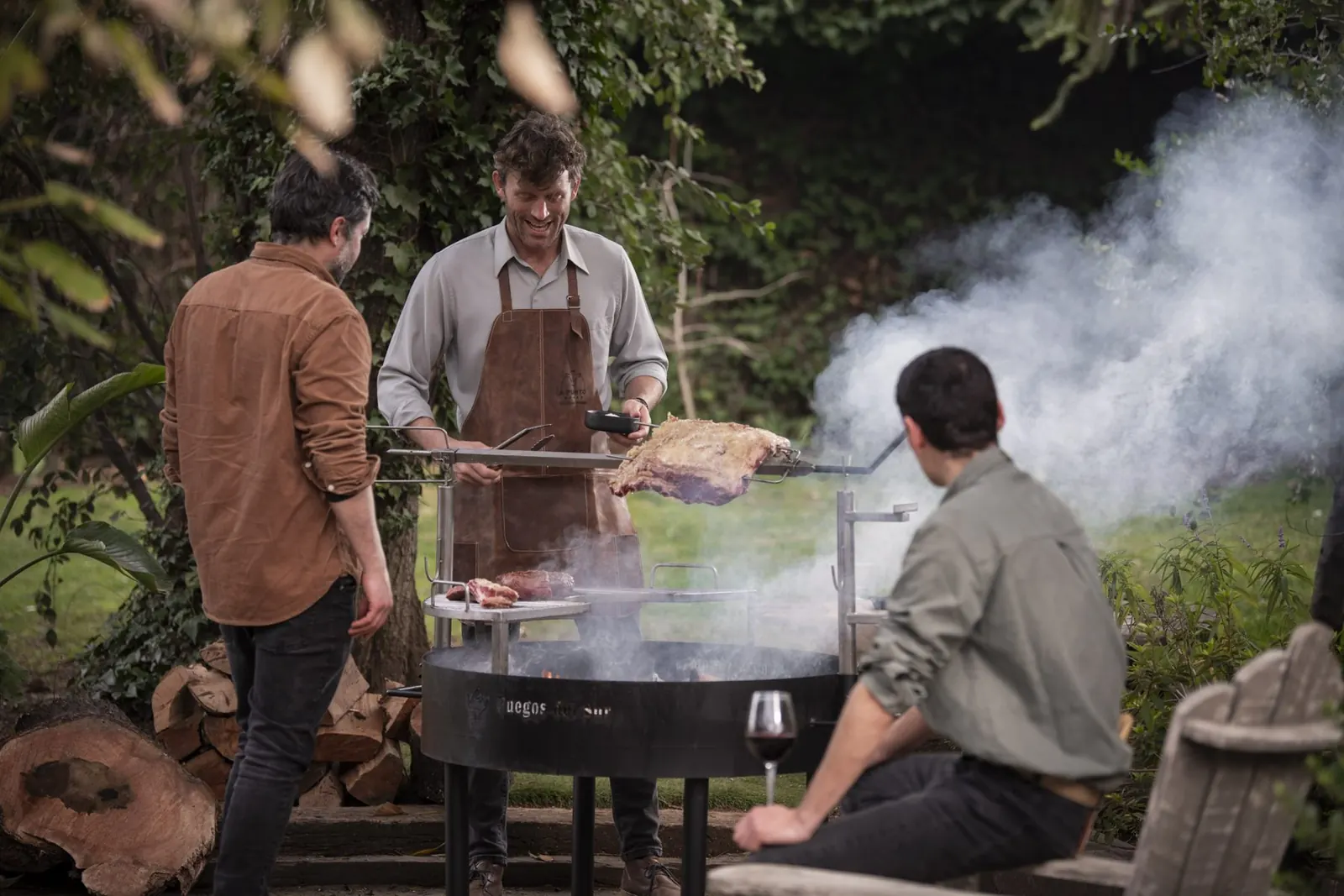 Maestro asador A Punto atendiendo una parrilla con humo en un jardín, junto a dos invitados